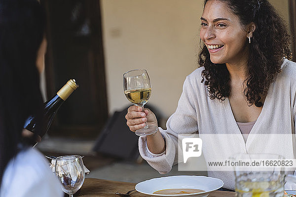 Lächelnde Frau hält ein Weinglas in der Hand und sieht einen Freund an  der am Esstisch im Hinterhof sitzt