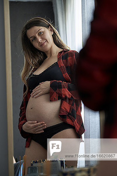 Lächelnde schwangere Frau mit Händen auf dem Bauch schaut in den Spiegel zu Hause