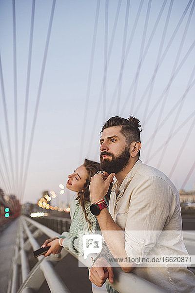 Nachdenkliches Paar  das sich bei Sonnenuntergang auf das Geländer einer Brücke in der Stadt stützt