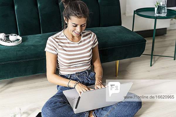 Lächelnde Geschäftsfrau  die einen Laptop benutzt und im Schneidersitz auf dem Boden vor dem Sofa im Büro sitzt