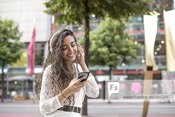 Lächelnde junge Frau  die Musik hört und ein Smartphone benutzt  während sie in der Stadt steht