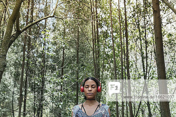 Junge Frau mit geschlossenen Augen hört Musik über Kopfhörer gegen Bäume im Park