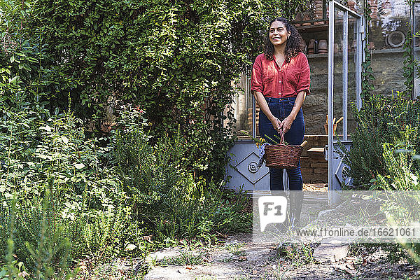Smiling mature woman looking away holding basket while standing in back yard