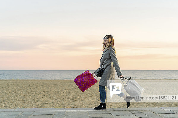 Lächelnde schöne Frau mit Einkaufstaschen  die auf der Promenade am Strand spazieren geht  während sie bei Sonnenuntergang gegen den Himmel schaut