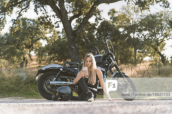 Blonde Motorradfahrerin  die ihr Smartphone benutzt  während sie auf dem Motorrad am Straßenrand sitzt