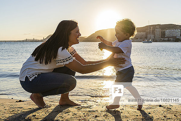 Fröhliche Mutter spielt mit ihrem Sohn am Strand bei Sonnenuntergang
