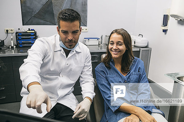 Ein männlicher Zahnarzt erklärt einer lächelnden jungen Patientin über einem Laptop in einer Klinik ein medizinisches Verfahren