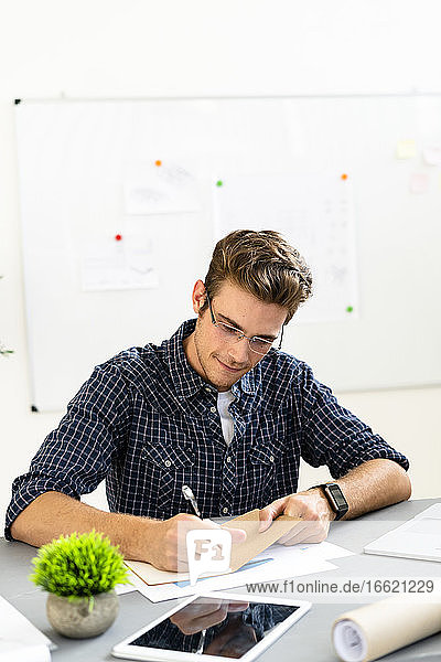 Architect writing on paper while sitting by desk at office