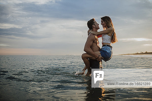 Man carrying woman while standing in water during sunset at beach
