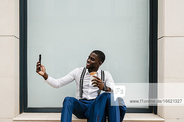 Lächelnde junge männliche Fachkraft  die ein Selfie mit ihrem Smartphone macht  während sie am Fenster in der Stadt sitzt