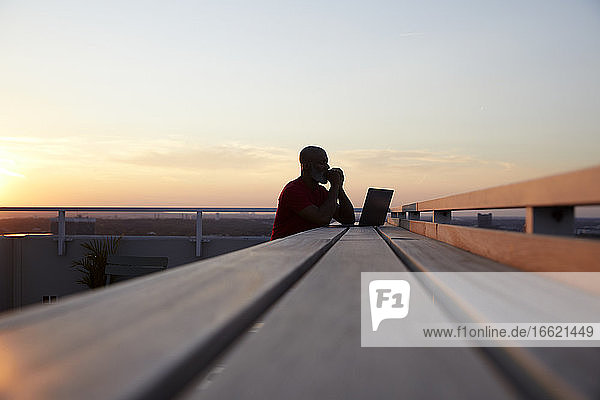 Surface level view of wooden table with man and laptop at rooftop against sky during sunset