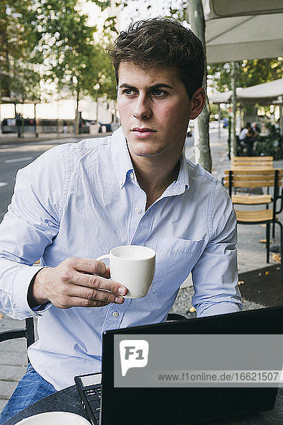 Nachdenklicher junger Mann hält eine Kaffeetasse  während er mit seinem Laptop in einem Straßencafé sitzt