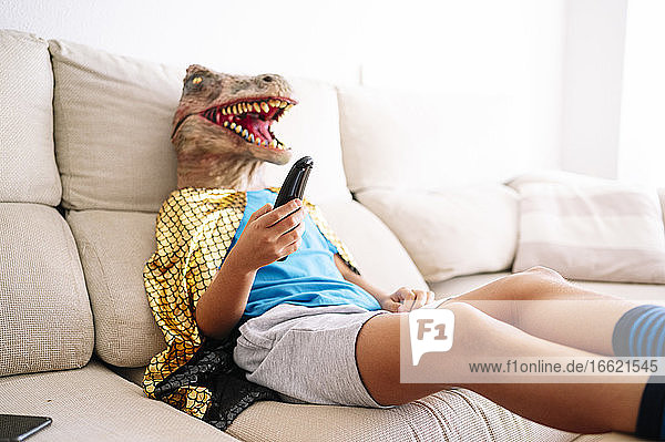 Junge mit Dinosauriermaske sieht fern und entspannt sich auf dem Sofa zu Hause