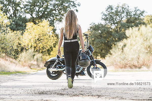 Blonde Frau hält einen Helm  während sie auf der Straße auf ein Motorrad zugeht