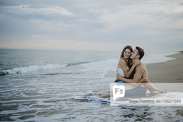 Junger Mann küsst Frau  die auf seinem Schoß am Strand sitzt