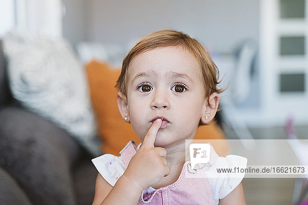 Thoughtful baby girl with finger on lips sitting on sofa at home