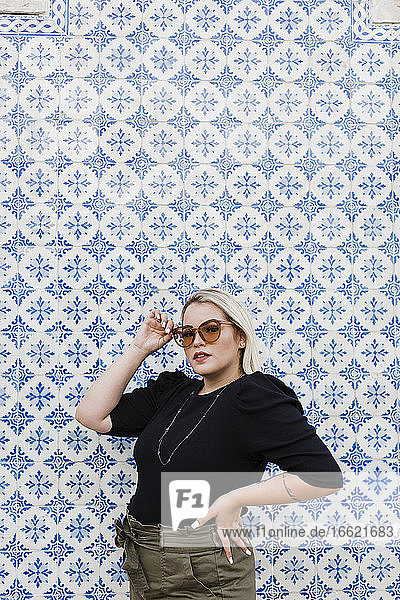 Stilvolle junge Frau mit Sonnenbrille  die an einer gekachelten Wand steht