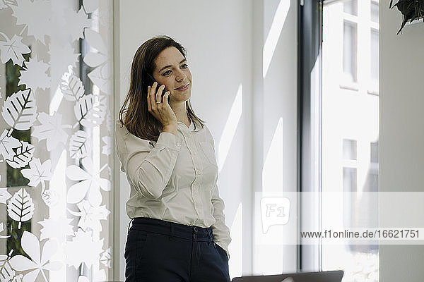 Geschäftsfrau mit Hand in der Tasche  die im Büro stehend mit ihrem Mobiltelefon spricht