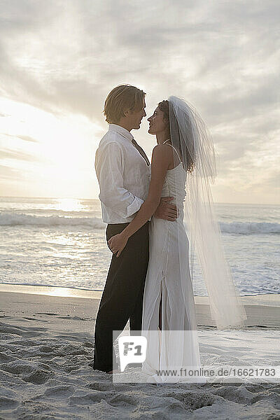 Neuvermähltes Paar  das sich bei Sonnenuntergang auf dem Sand stehend ansieht
