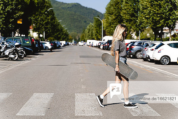 Junge blonde Frau hält ein Skateboard  während sie über einen Zebrastreifen in der Stadt läuft