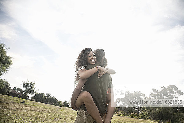 Junge Frau umarmt Mann  der sie in den Armen trägt  während sie in einem öffentlichen Park steht