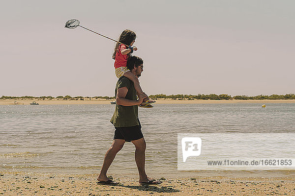 Vater trägt Tochter auf den Schultern bei einem Strandspaziergang gegen den klaren Himmel an einem sonnigen Tag