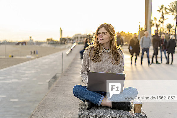 Thoughtful young woman looking away while sitting cross-legged with laptop and disposable cup on bench at promenade