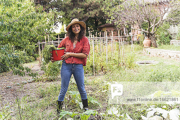 Lächelnde schöne Frau hält geerntetes Gemüse in einem Eimer im Garten