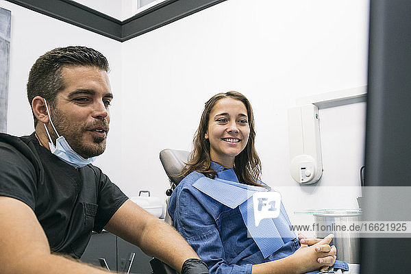 Gut aussehender männlicher Zahnarzt  der einer lächelnden Patientin über einem Laptop in einer Klinik ein medizinisches Verfahren erklärt