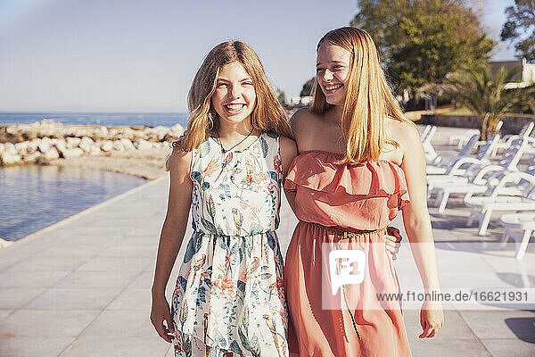 Smiling girls standing on sidewalk against sea during sunny day