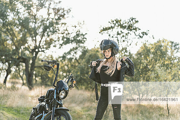 Blonde junge Bikerin mit Lederjacke  die neben einem Motorrad steht
