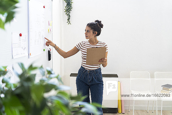Young businesswoman pointing at whiteboard while holding clipboard in creative workplace