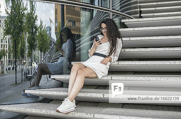 Junge Frau mit langen Haaren benutzt ein Mobiltelefon  während sie auf einer Treppe in der Stadt sitzt