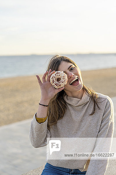 Fröhliche junge Frau hält sich einen frischen Donut vor das Gesicht  während sie bei Sonnenuntergang am Strand sitzt