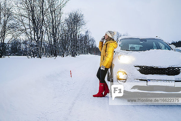 Ältere Frau spricht über Handy  während sie neben einem Auto in einer verschneiten Landschaft steht