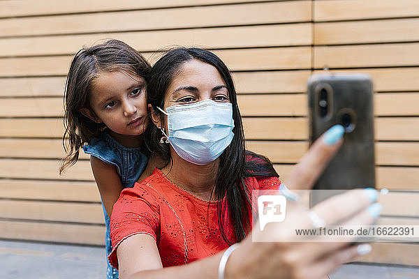 Mutter mit Maske macht Selfie mit Tochter an der Wand