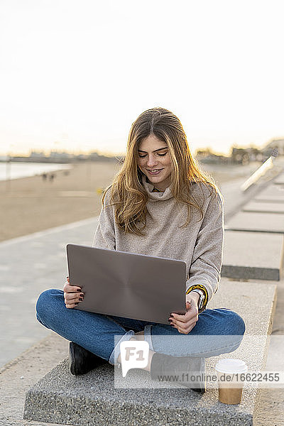 Lächelnde junge Frau sitzt im Schneidersitz mit Laptop und Einwegbecher auf einer Bank an der Promenade gegen den Himmel