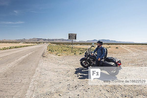 Man sitting on motorcycle parked by desert road against sky during road trip  Nevada  USA