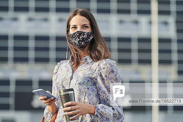 Junge Frau mit Gesichtsmaske  die ein Mobiltelefon benutzt  während sie in der Stadt steht