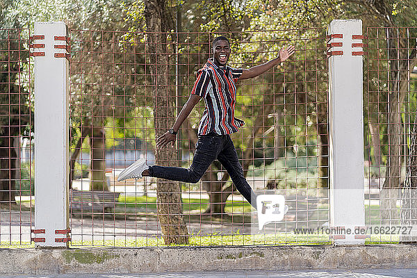 Cheerful young man jumping against fence at park
