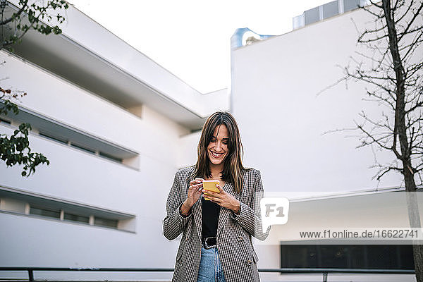 Lächelnde Frau  die eine Textnachricht auf ihrem Smartphone sendet  während sie an der Außenseite eines Gebäudes steht