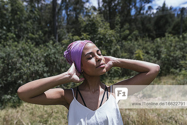 Junge Frau trägt lila Kopftuch  während sie mit geschlossenen Augen im Park an einem sonnigen Tag steht