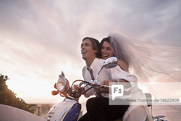 Fröhliches frisch vermähltes Paar im Hochzeitskleid fährt Motorroller bei Sonnenuntergang