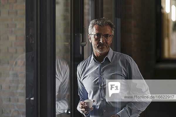 Geschäftsmann mit Brille  der eine Kaffeetasse hält und sich im Büro an ein Fenster lehnt