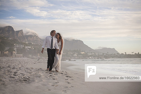 Glückliches frisch verheiratetes Paar spaziert am Strand gegen den Himmel bei Sonnenuntergang