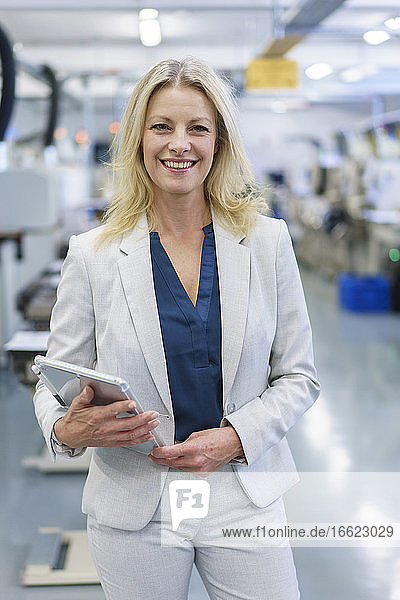Lächelnde reife blonde Geschäftsfrau  die ein digitales Tablet hält  während sie vor einer beleuchteten Industrie steht