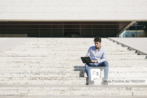 Selbstbewusster junger Mann  der einen Laptop benutzt  während er an einem sonnigen Tag auf einer Treppe sitzt