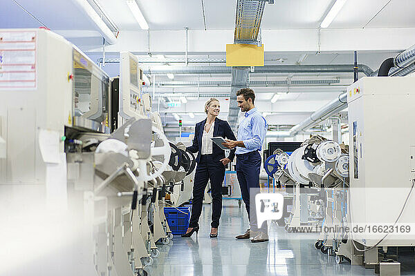 Ein männlicher Techniker hält ein digitales Tablet in der Hand  während er sich mit einer Geschäftsfrau unterhält  die an einer Maschine in einer Fabrik steht.