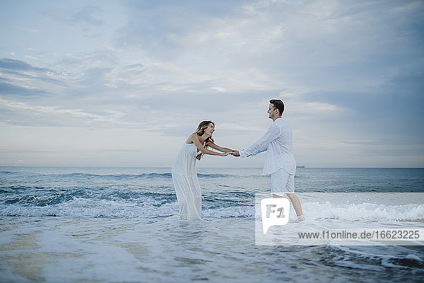 Verspieltes Paar hält sich an den Händen und steht im Wasser am Strand