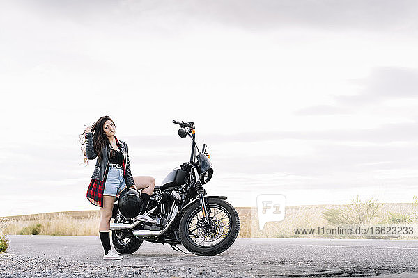 Young female biker standing on road with motorbike